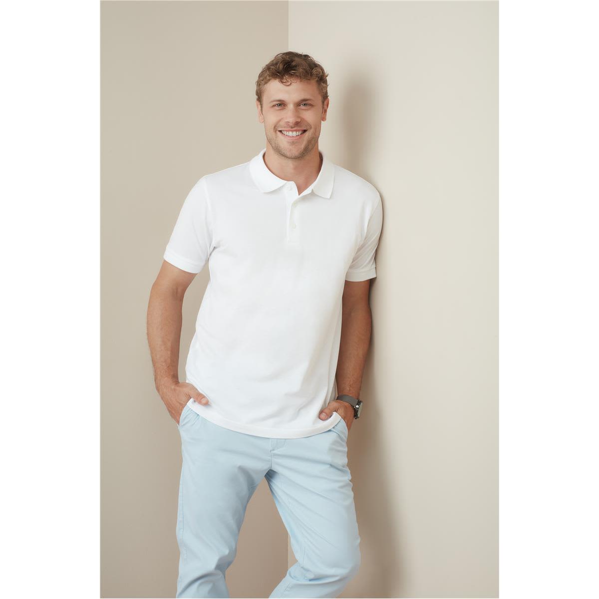 A man wearing a white premium cotton polo shirt, smiling while leaning against a wall, paired with light blue trousers. Men's Premium Cotton Polo - Image 1