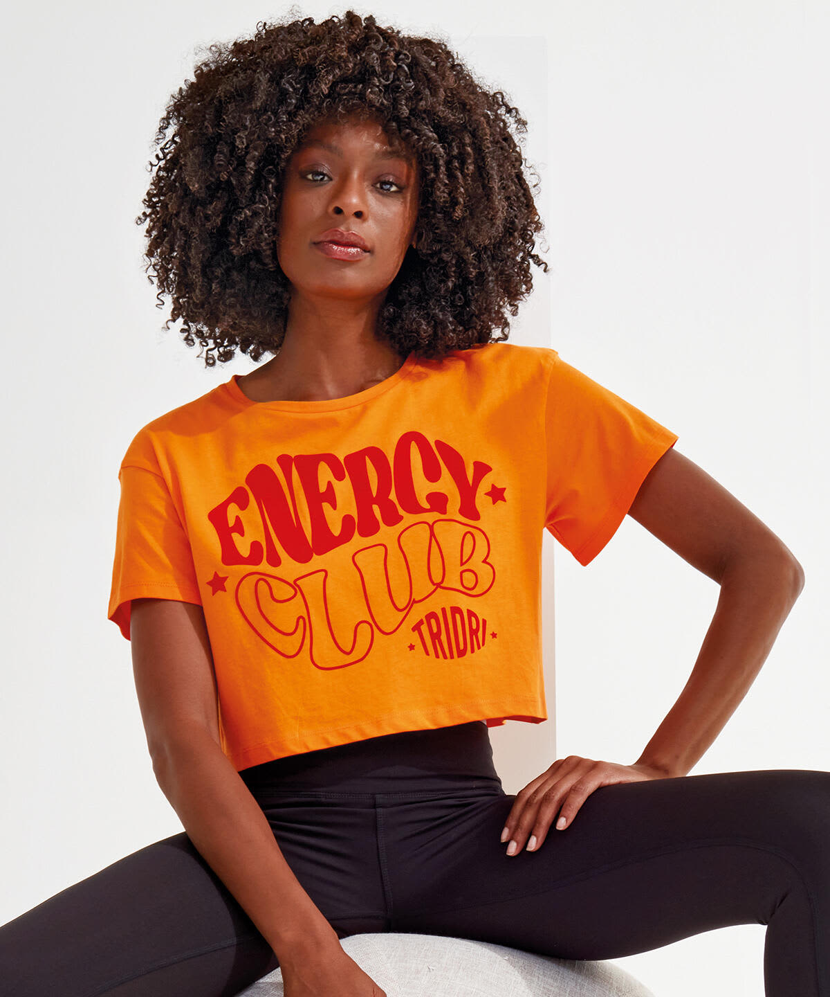 A model wearing an orange crop top with red text, featuring a logo, paired with black leggings. Crop Top - Image 1