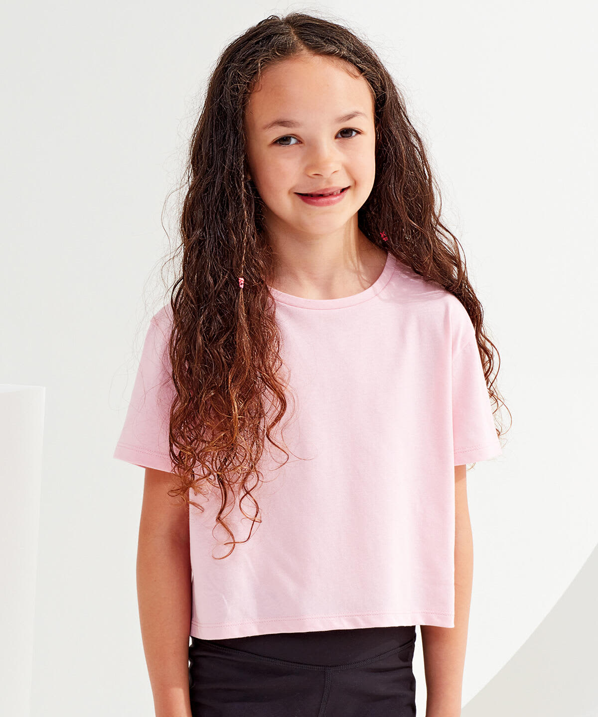 A pink kids boxy T-shirt on a girl with long, curly hair, standing against a light background. Kids Boxy T-Shirt - Image 1
