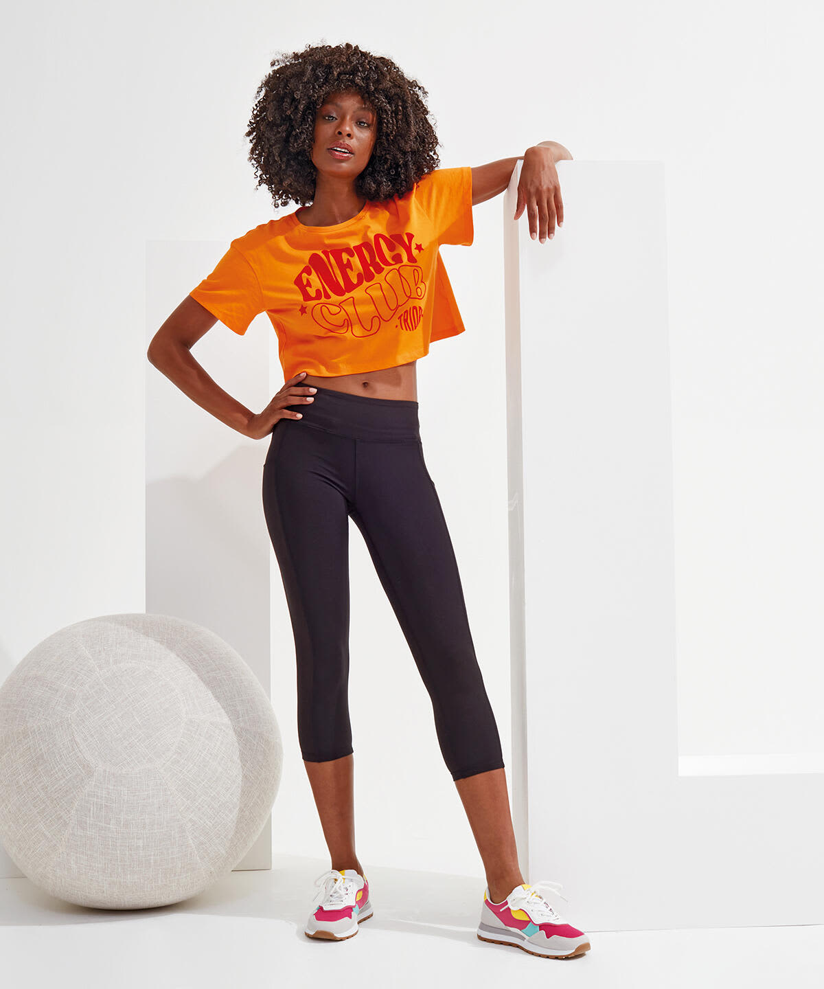 Black 3/4 length leggings displayed on a model, paired with an orange cropped t-shirt and colourful sneakers. Recycled Performance Leggings 3/4 Length - Image 1