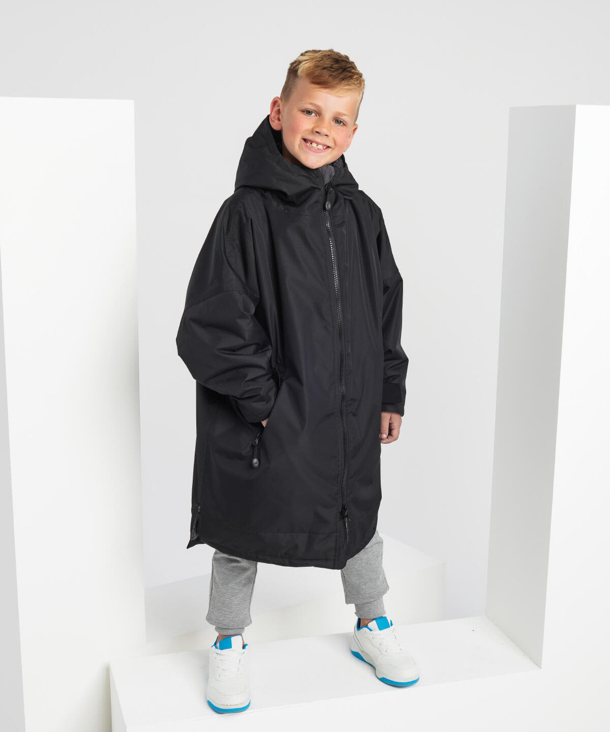 A black waterproof changing robe with a hood, worn by a child standing on a white display. Kids All-Seasons Waterproof Changing Robe - Image 1