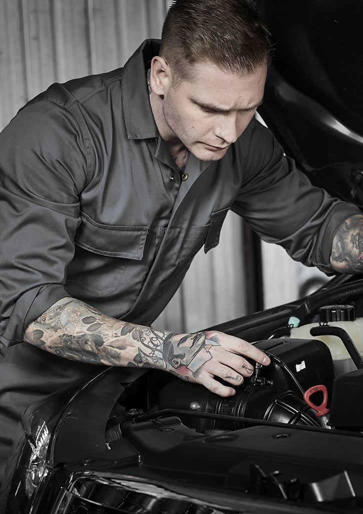 A man in a grey service overall works on a car engine, showcasing detailed tattoos on his arms. The image features a logo on the overall. Mens Service Overall - Image 1
