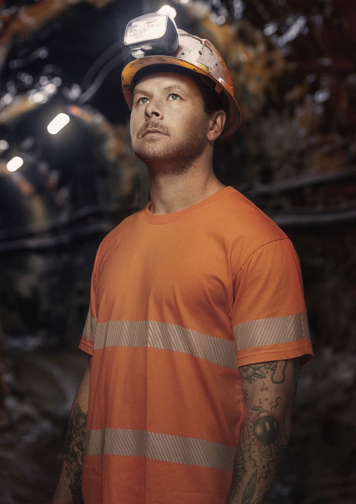 A man wearing an orange hi-vis segmented tape cotton tee shirt and a safety helmet with headlamp, standing in a dimly lit area. Mens Hi Vis Segmented Tape Cotton Tee - Image 1