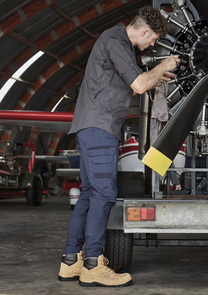 Mens Streetworx Heritage Pant in navy, paired with tan work boots, worn by a man repairing machinery in a hangar. Mens Streetworx Heritage Pant - Cuffed - Image 1