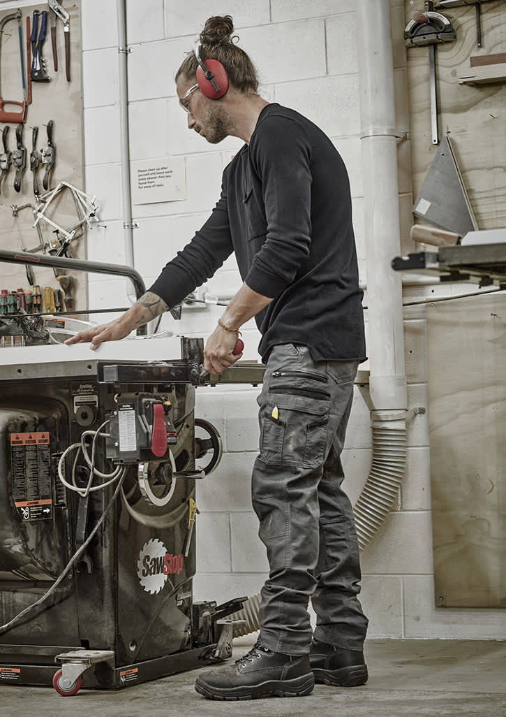 A man wearing black work pants stands by a workshop table, handling tools in a well-equipped space. Mens Streetworx Heritage Pant - Image 1