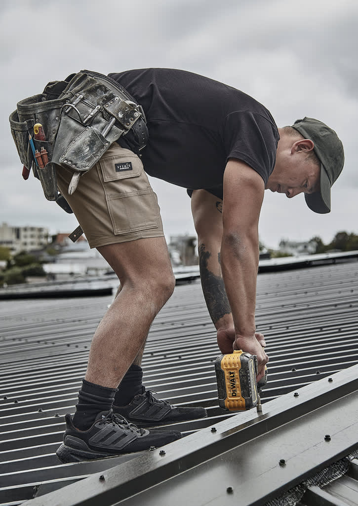 A person wearing khaki cargo shorts and a black t-shirt is using a power drill on a roof. Tools hang from a tool belt. Mens Essential Stretch Cargo Short - Image 1