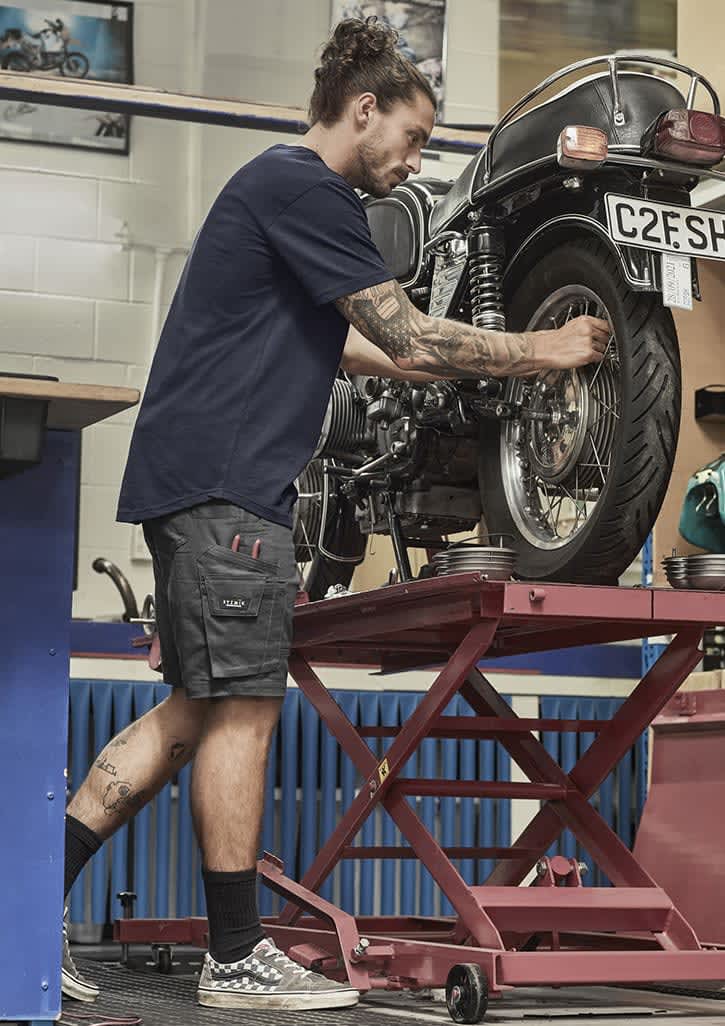 A man in navy blue shorts works on a motorcycle, showcasing rugged cooling stretch cargo shorts with pockets. Mens Rugged Cooling Stretch Short - Image 1