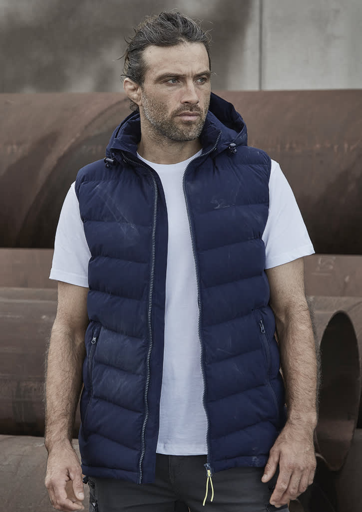 A man wearing a navy blue unisex hooded puffer vest over a white t-shirt, standing in an industrial setting. Unisex Streetworx Hooded Puffer Vest - Image 1
