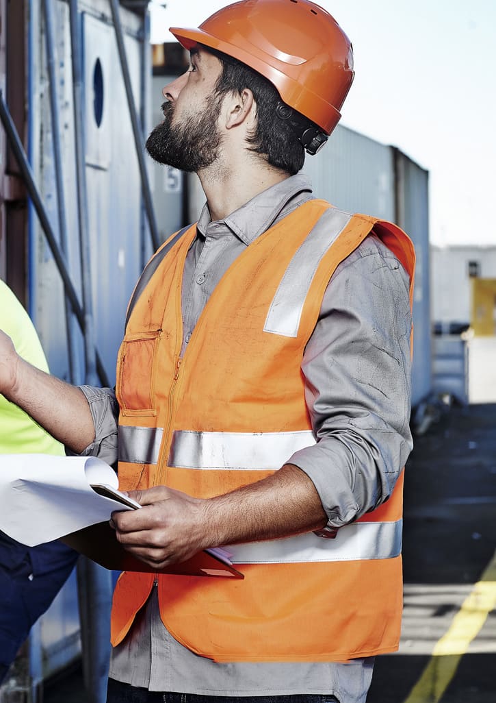 A man wearing an orange hi-vis zip vest with reflective stripes and a hard hat, holding documents at a construction site. Unisex Hi Vis Zip Vest - Image 1