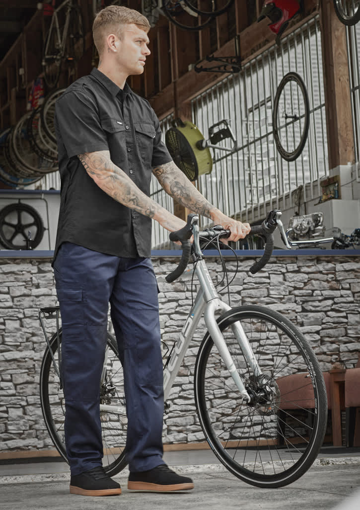 A person wearing navy midweight drill cargo pants and a black shirt stands beside a bicycle in a workshop setting. Mens Midweight Drill Cargo Pant (Stout) - Image 1