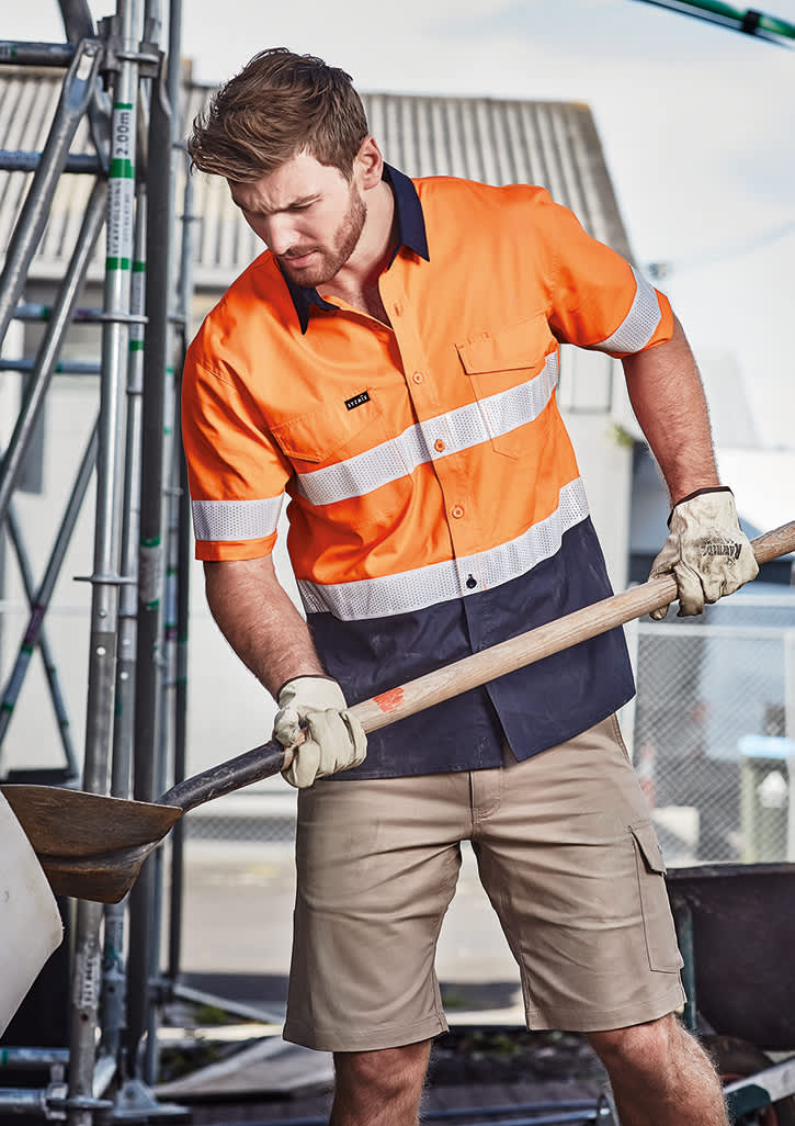 A short sleeve work shirt in orange and navy with reflective tape, worn by a man using a shovel, with a construction setting in the background. Mens Rugged Cooling Hi Vis Taped Short Sleeve Shirt - Image 1