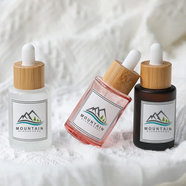 A collection of three 30mL frosted glass dropper bottles in clear, pink, and dark amber with bamboo lids and logos. 30mL Frosted Glass Dropper Bottle - Image 1