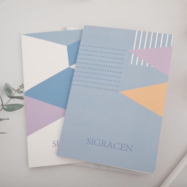 A set of two A5 soft cover ruled notebooks featuring geometric designs in blue, white, purple, and orange, with a logo. 2 Pack A5 Soft Cover Ruled Notebook Set - Image 1