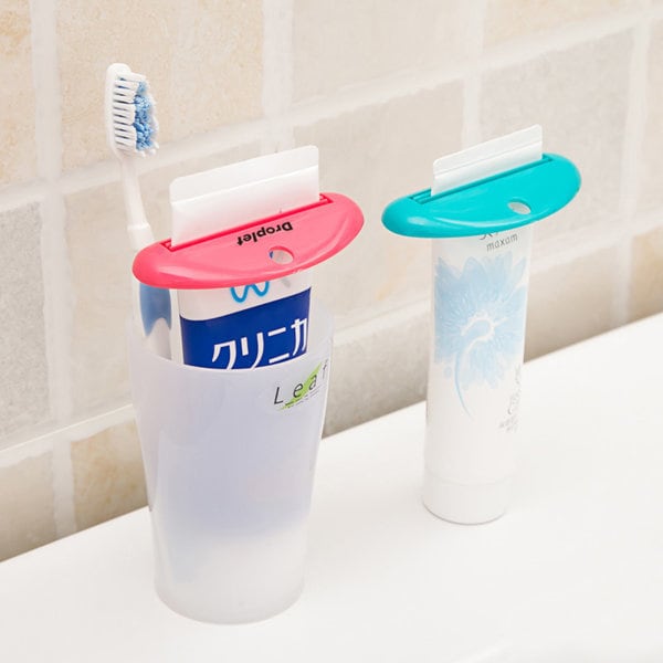 A toothpaste tube squeezer dispenser in pink and blue, alongside a toothbrush, in a bathroom setting. Toothpaste Tube Squeezer Dispenser - Image 1