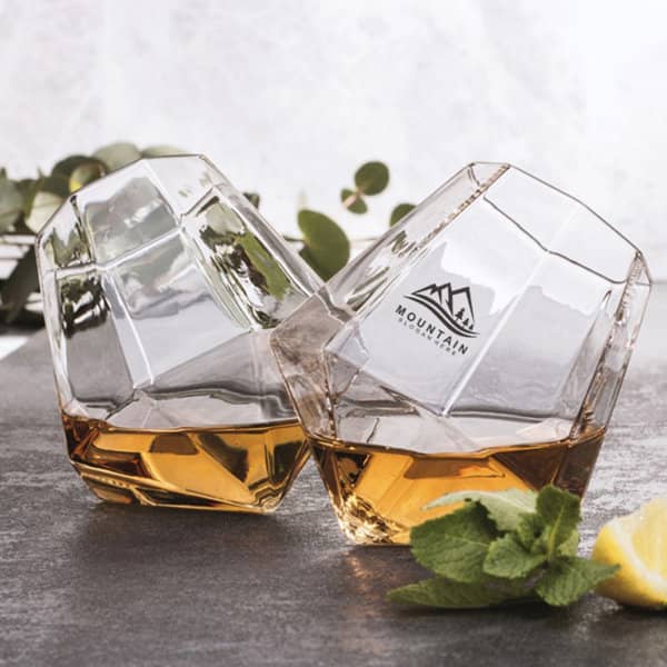 Two diamond-shaped stemless wine glasses in clear glass, featuring a logo, filled with a brown liquid. Promotional Diamond Shaped Stemless Wine Glass - Image 1