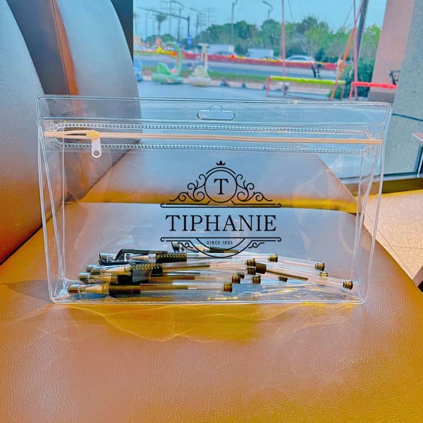 A clear PVC zipper bag containing various small metal items, with a decorative logo on the front. PVC Zipper Bag - Image 1