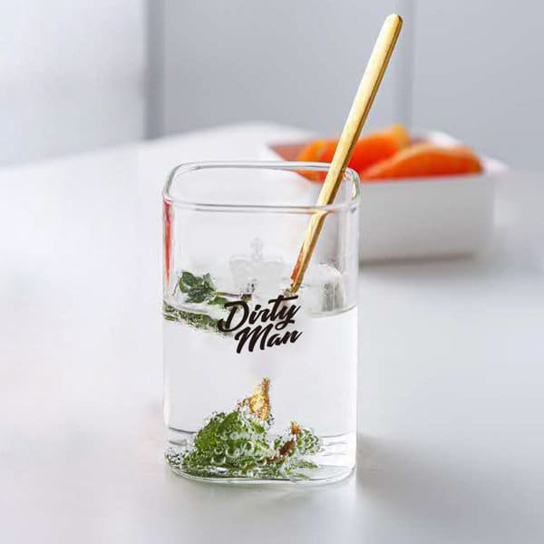 A square 9 oz. clear glass water cup filled with water and mint leaves, featuring a logo and a gold stirring stick. 9 oz. Square Glass Water Cup - Image 1