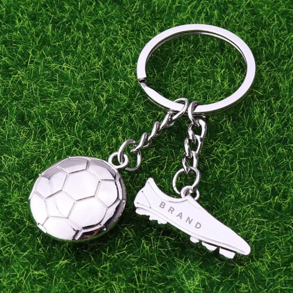 A silver football charm keychain with a soccer ball and a boot, featuring a branding logo. Football Charm Keychain - Image 1