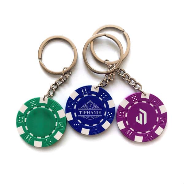 Three poker chip keychains in green, blue, and purple, attached to silver key rings, each featuring a logo. Poker Chip Keychain - Image 1