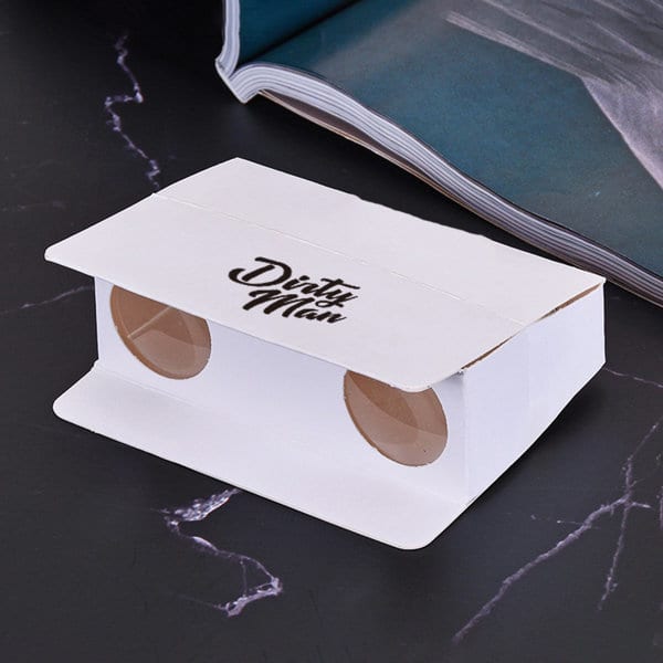 A custom folding paper binocular in white with circular lenses and a logo. The background features a magazine. Custom Folding Paper Binocular - Image 1