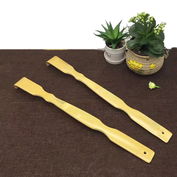 A bamboo back scratcher in a light yellow colour, featuring a curved design, placed on a brown surface with small plants. Bamboo Back Scratcher - Image 1