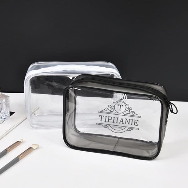 Two large clear PVC zippered toiletry bags in black and transparent, featuring a logo. Makeup brushes are placed beside them. Large Clear PVC Zippered Toiletry Bag - Image 1