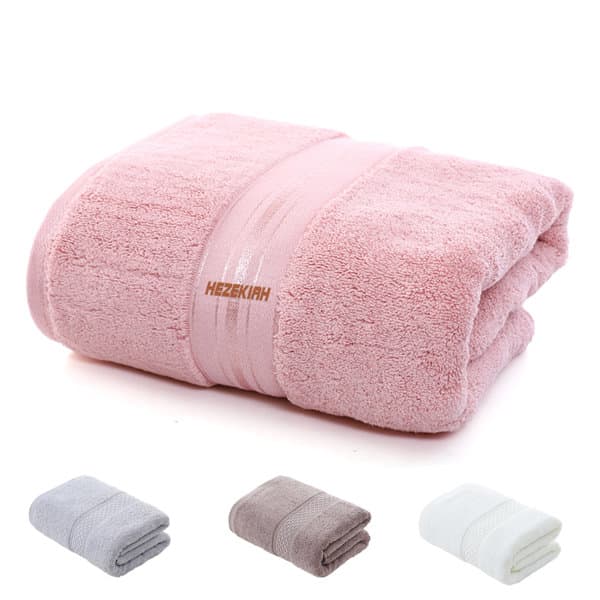 A pink extra large cotton bath towel is neatly folded, with grey, brown, and white towels folded beside it. The towel has a logo. Extra Large Cotton Bath Towel - Image 1
