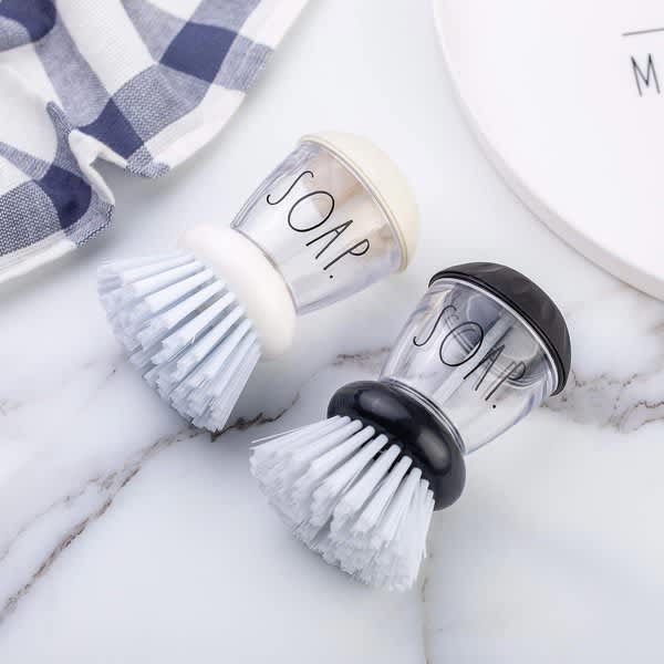 A small soap dispensing palm brush in white and black, featuring bristles and a transparent soap chamber. Small Soap Dispensing Palm Brush - Image 1