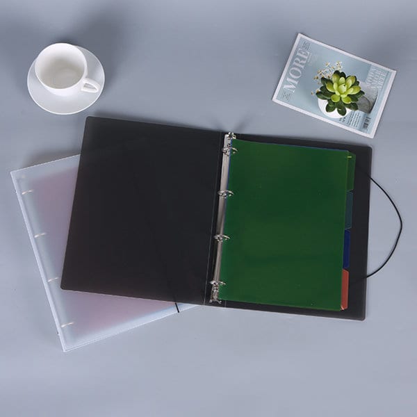 A4 frosted transparent ring binder in black with green and blue dividers, placed on a grey surface with a cup and a magazine. A4 Frosted Transparent Ring Binder - Image 1