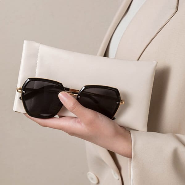 A hand holds a black pair of sunglasses alongside a cream-coloured storage bag. The bag features a logo. Shrapnel Sunglasses Storage Bag - Image 1