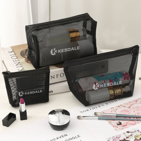 Three black mesh cosmetic bags of varying sizes displayed on a table with beauty products and a magazine. Mesh Cosmetic Bag Set - Image 1