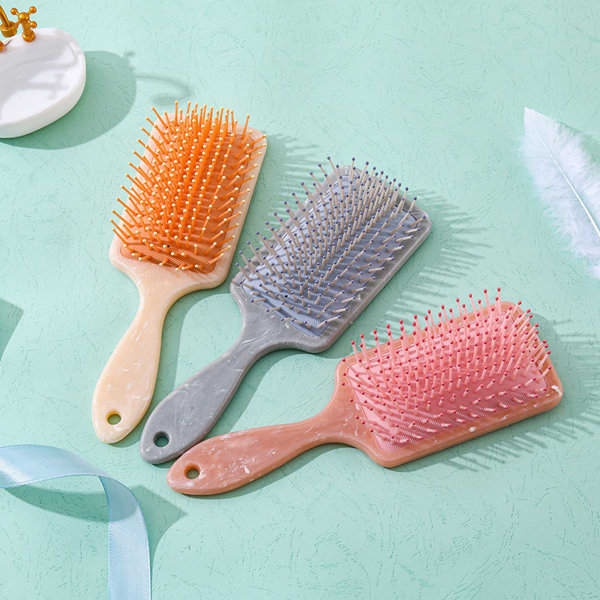 Three Paddle Detangler Hair Brushes in peach, grey, and pink on a light green surface, featuring branding on the handles. Paddle Detangler Hair Brush - Image 1