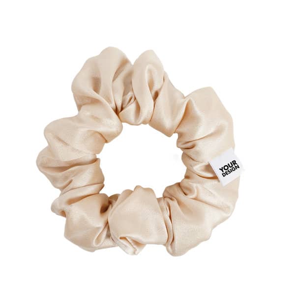 A beige satin scrunchie featuring a small tag with branding. Soft and luxurious texture showcased in a round shape. Custom Satin Scrunchie - Image 1