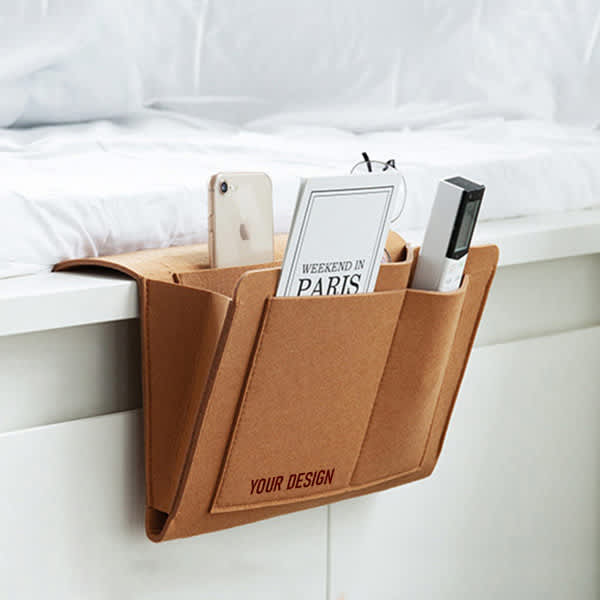 A tan felt bedside pocket with multiple compartments holding a phone, a book, and a remote control. Felt Bedside Pocket - Image 1