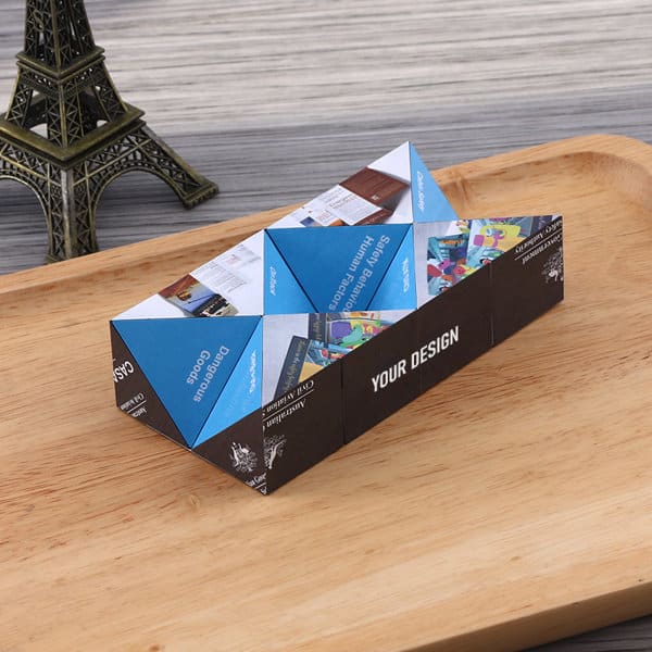 A fold-out magic cube in blue and black, displaying various designs on its triangular panels, placed on a wooden tray. Promotional Fold Out Magic Cube - Image 1