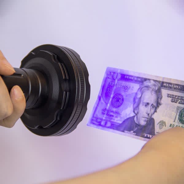 A UV LED flashlight in black is illuminating a banknote, highlighting its security features. UV LED Flashlight - Image 1