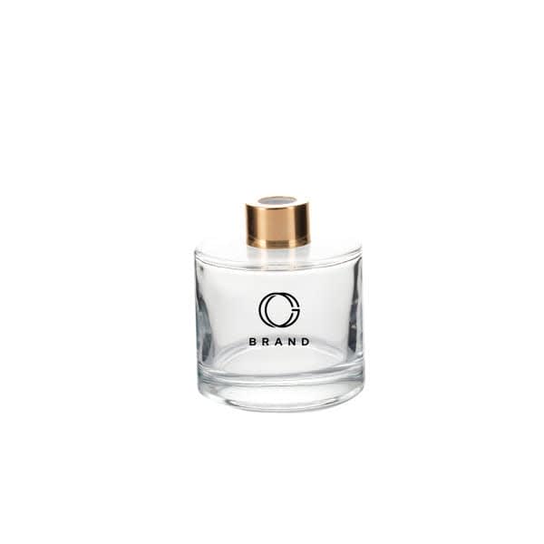 A round glass diffuser bottle in clear with a gold cap, featuring a logo on the front. 5 oz. Diffuser Round Glass Bottle With Gold Cap - Image 1