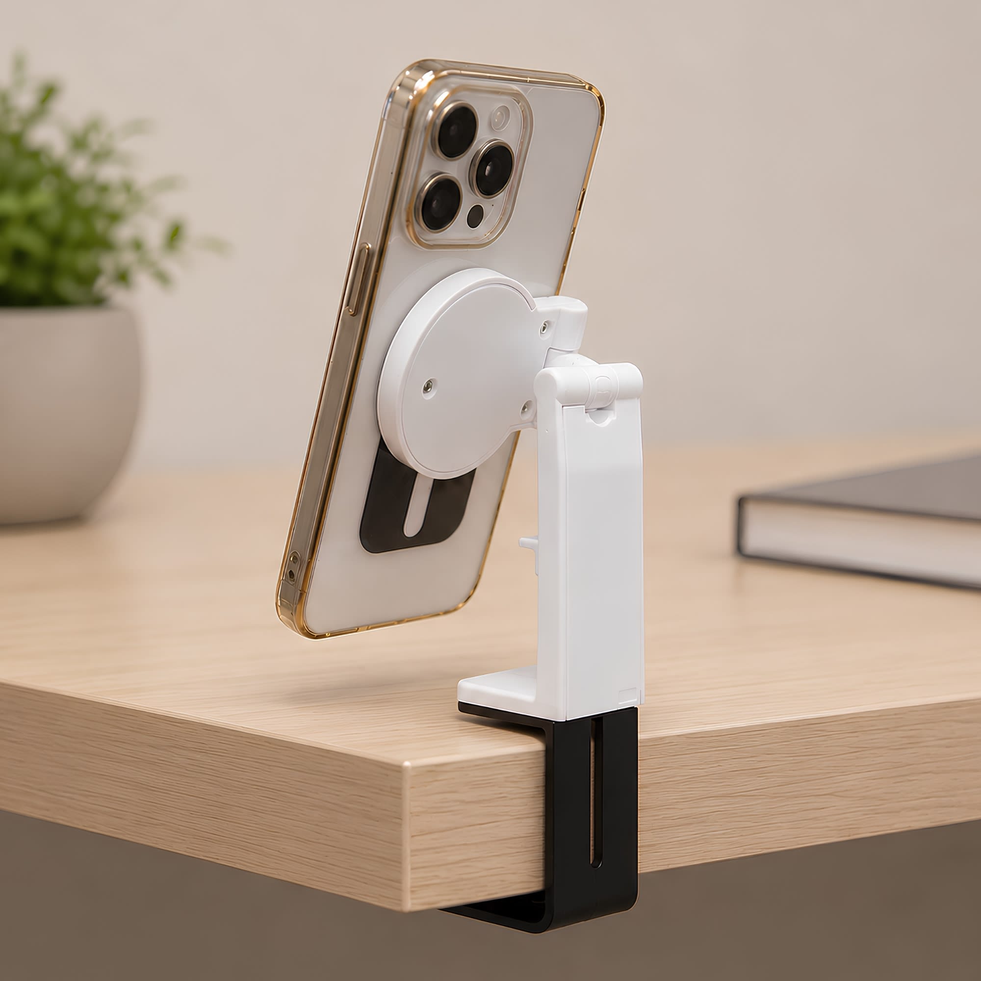 A white and black mobile phone stand holds a phone, attached to a wooden desk. A small plant and a book are in the background. Traveler Mobile Phone Stand - Image 1