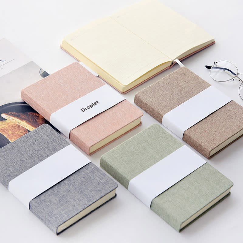 A collection of classic cloth bound notebooks in grey, pink, beige, and green, with a white band around them. Classic Cloth Bound Notebook - Image 1